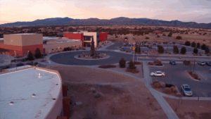 Aerial view of a modern building complex with a circular driveway, parking lots, and surrounding desert landscape at sunset, with mountains in the background.