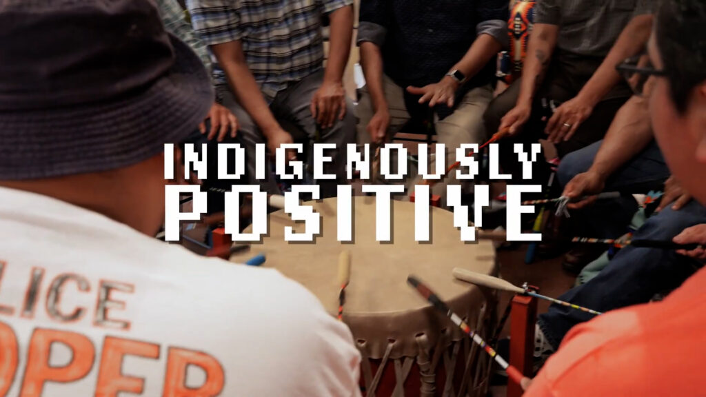 A group of people play a large hand drum together in a circle. Bold text over the image reads, "Indigenously Positive.