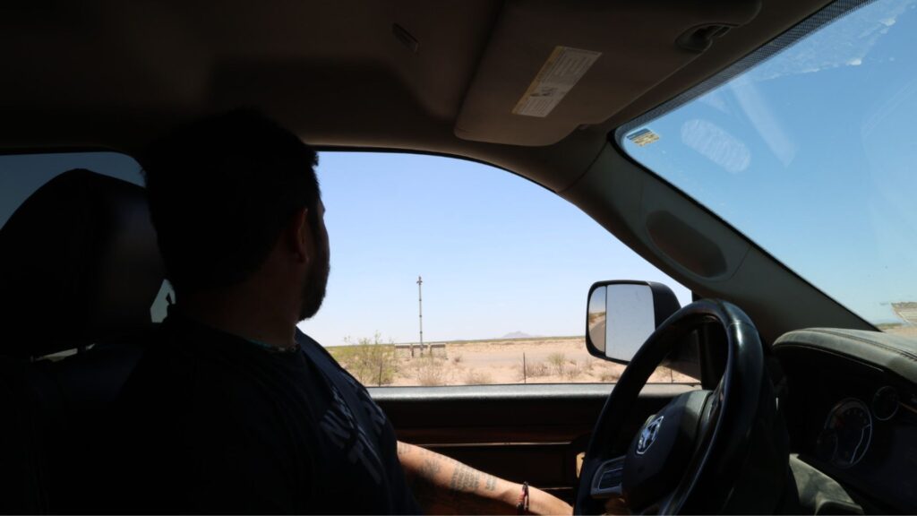 A person sits in the driver's seat of a vehicle parked in a desert area, looking out the window at the dry landscape and clear sky.