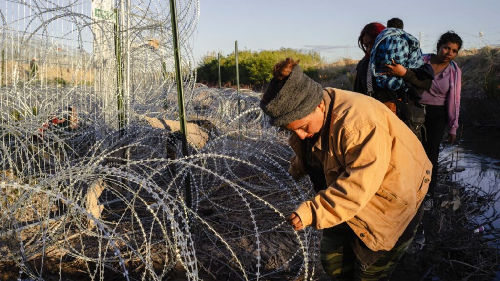 A woman bends over razor wire at a border fence while others, including a person carrying a child, stand nearby in an outdoor setting.