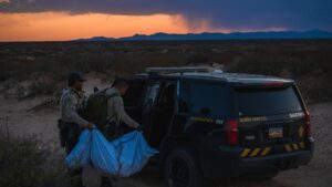 Two sheriff deputies stand by a patrol SUV in a desert landscape at dusk, one holding a large white bag as they interact near the vehicle.