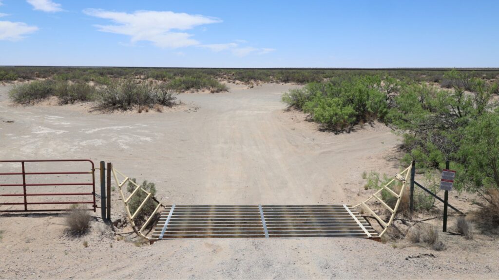 A metal cattle guard spans a dirt road in a dry, desert landscape with sparse bushes and a red gate to the left.