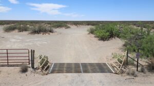 A metal cattle guard spans a dirt road in a dry, desert landscape with sparse bushes and a red gate to the left.