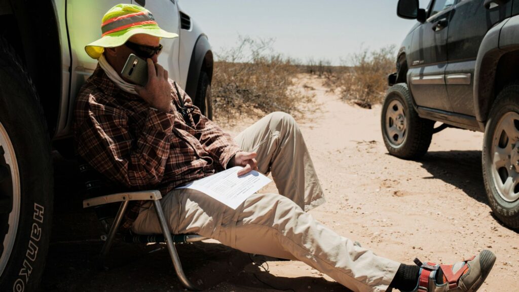 A person in outdoor clothing sits on a folding chair between two vehicles in a desert, talking on a phone and holding papers.