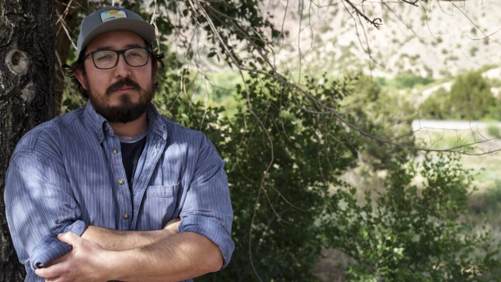 A man with a beard and glasses, wearing a cap and a blue shirt, stands outdoors with his arms crossed in front of leafy trees.