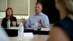A man in a blue shirt speaks at a meeting table with water bottles, seated next to a woman with red hair. Nameplates are visible in front of them.