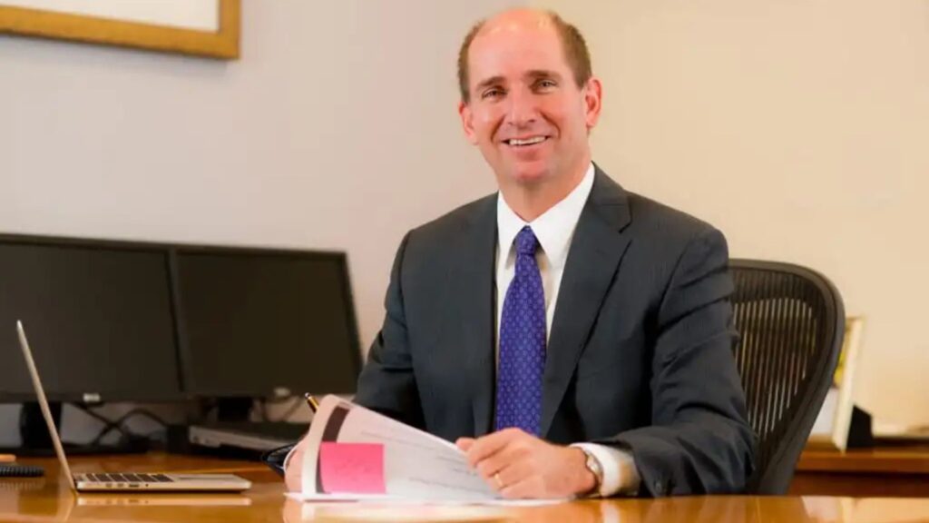 A man in a business suit sits at a desk with a laptop, holding documents and smiling at the camera. Two monitors and framed art are in the background.