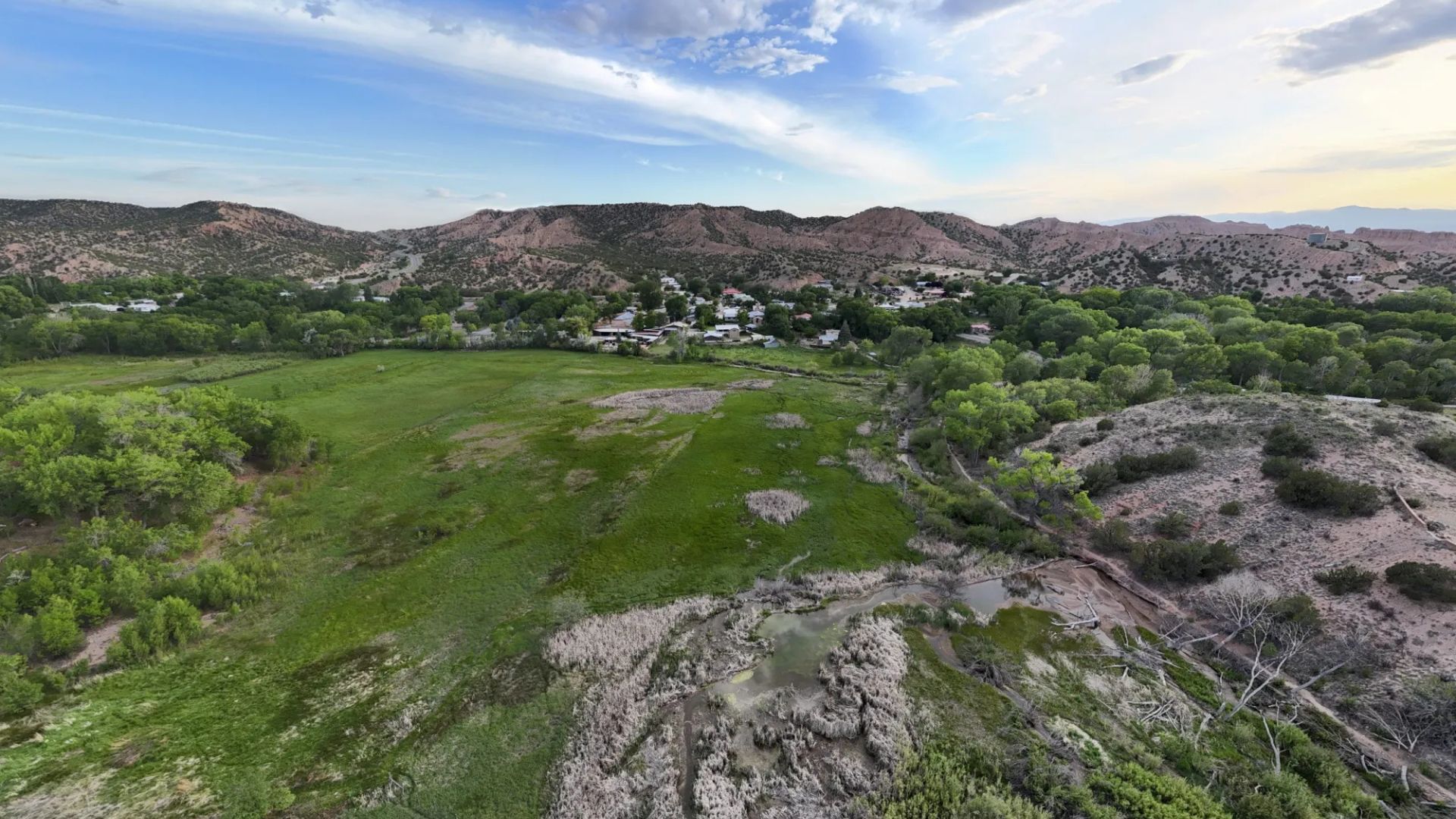 Aerial view of a small town surrounded by green fields, trees, and hills under a partly cloudy sky.