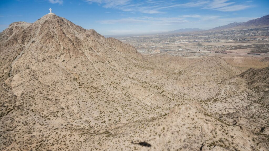 A rocky desert mountain landscape with a large white cross at the peak, overlooking a city in the background under a partly cloudy sky.