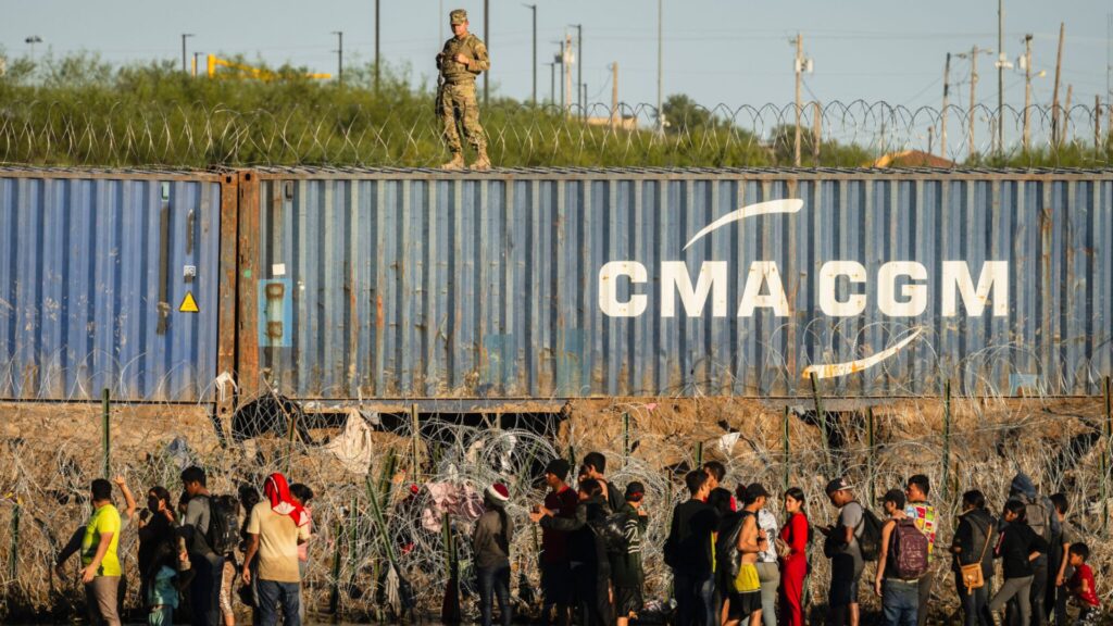 A group of people stand near a fence covered in razor wire, while a uniformed guard stands atop a large shipping container marked "CMA CGM.