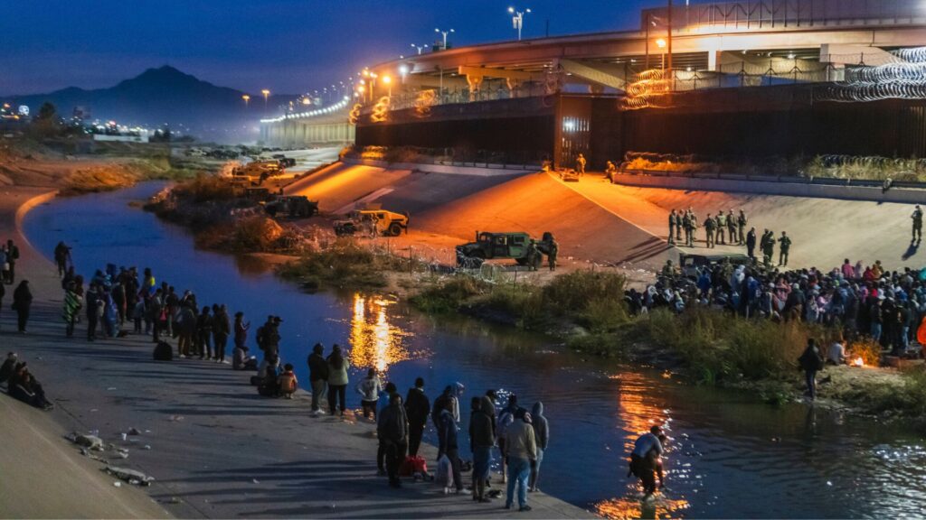 A group of people gather along a concrete riverbank at dusk, with vehicles and officials standing nearby under an overpass with barbed wire.