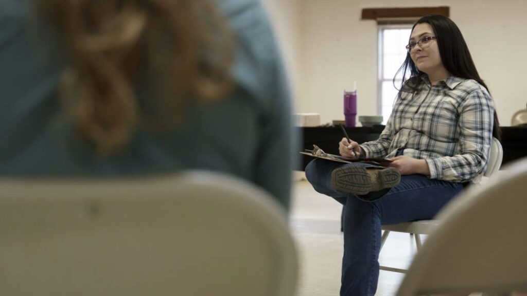 A woman in a plaid shirt sits on a chair with a notebook and pen, listening attentively in a room with another person in the foreground.