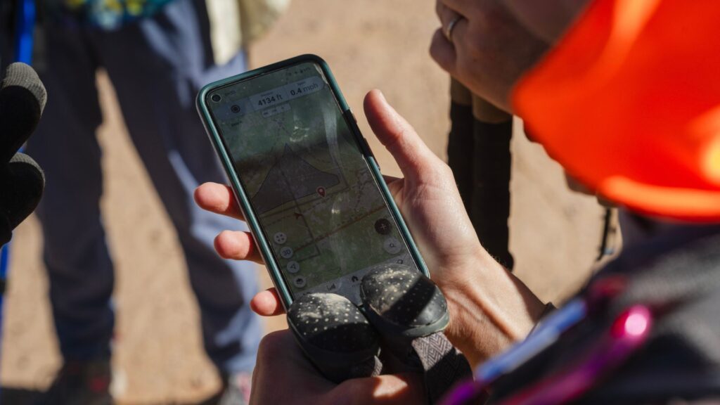 A person holding trekking poles checks a map app on their smartphone while standing outdoors on a dirt surface.