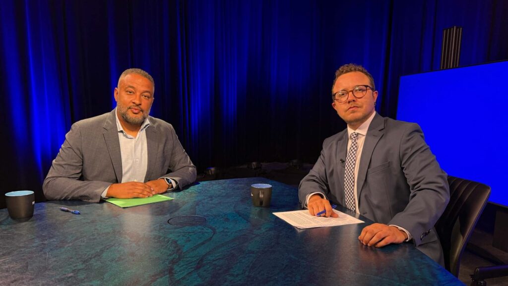 Two men in suits sit at a round table with papers and cups, facing the camera, against a dark blue studio background.