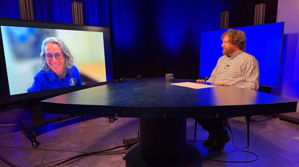A man sits at a round table in a studio, interviewing a woman who appears on a large screen. The background is lit with blue lighting.