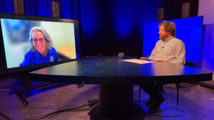 A man sits at a round table in a studio, interviewing a woman who appears on a large screen. The background is lit with blue lighting.