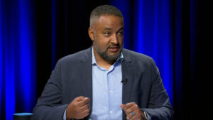 A man in a suit jacket and light blue shirt speaks while gesturing with his hands. He is seated indoors against a dark background with blue lighting.