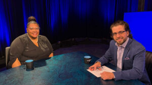 Two people sit at a table with mugs and papers in a studio setting with blue lighting, preparing for or engaged in a discussion or interview.