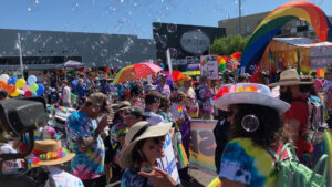 A crowd of people in colorful tie-dye shirts and hats gathers at an outdoor parade with rainbow decorations, balloons, and bubbles in the air.