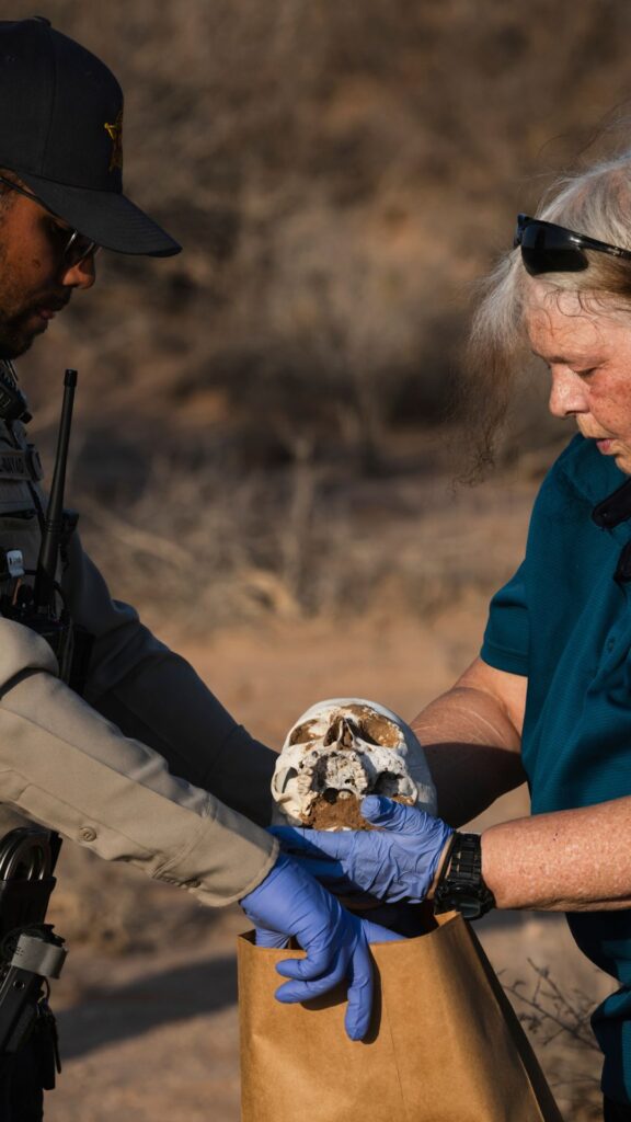 Two people wearing gloves carefully place a human skull into a brown paper evidence bag in an outdoor, dry setting.