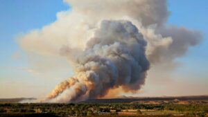 A large plume of smoke rises into the sky from a wildfire burning in a rural landscape with trees, open land, and distant hills.