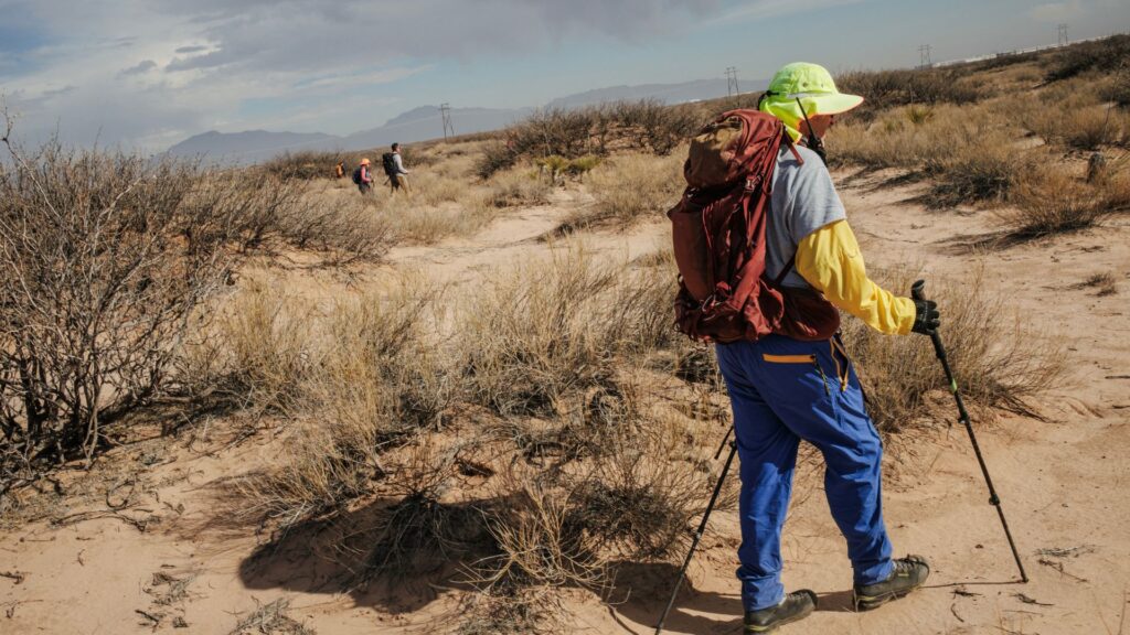 A person wearing hiking gear and using trekking poles walks through a dry, desert landscape with three people visible in the background.