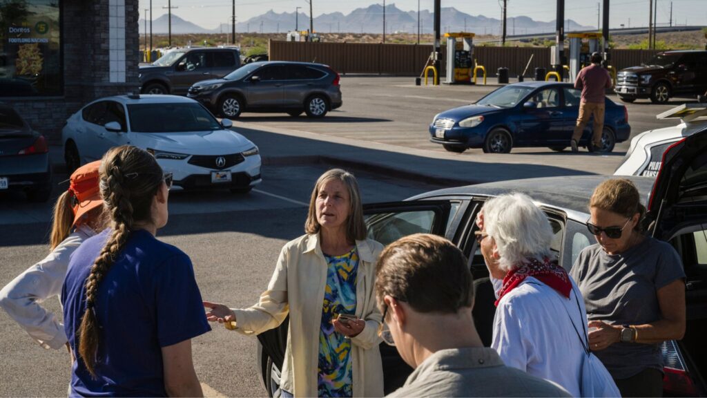 A group of people stand and talk in a sunny parking lot near parked cars, with gas pumps and mountains visible in the background.
