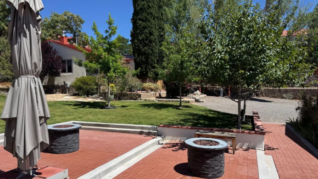 A backyard with red brick patio, two covered fire pits, green lawn, several trees, a gravel area, and a neighboring house in the background.