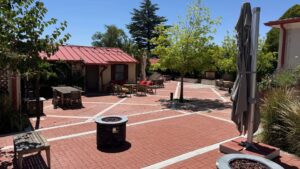 Brick patio with fire pit, outdoor seating, tables, chairs, closed umbrellas, and trees providing shade on a sunny day.