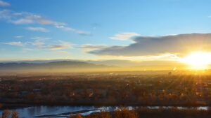 The sun rises over a cityscape with mountains in the distance, casting light over trees, buildings, and a river in the foreground.