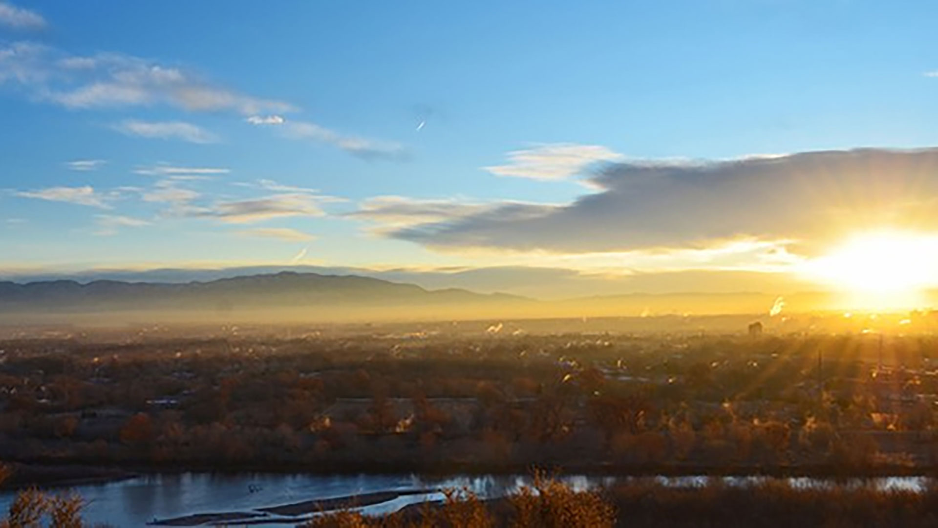The sun rises over a cityscape with mountains in the distance, casting light over trees, buildings, and a river in the foreground.