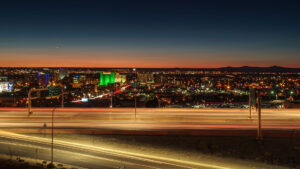 Nighttime cityscape with illuminated buildings and a highway in the foreground, showing light trails from passing vehicles, under a darkening sky.