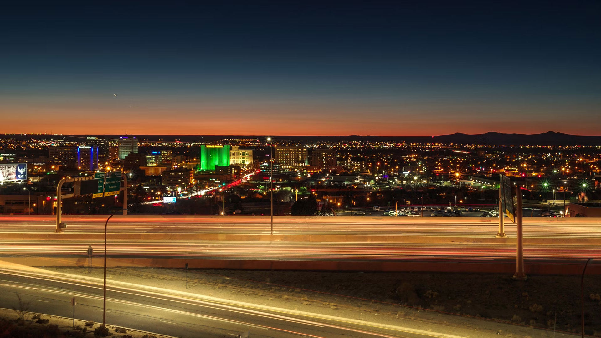 Nighttime cityscape with illuminated buildings and a highway in the foreground, showing light trails from passing vehicles, under a darkening sky.