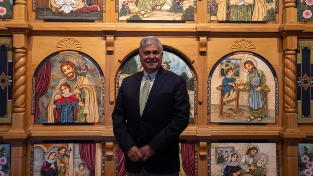 A man in a suit stands in front of a wooden wall decorated with colorful religious paintings depicting biblical scenes and figures.