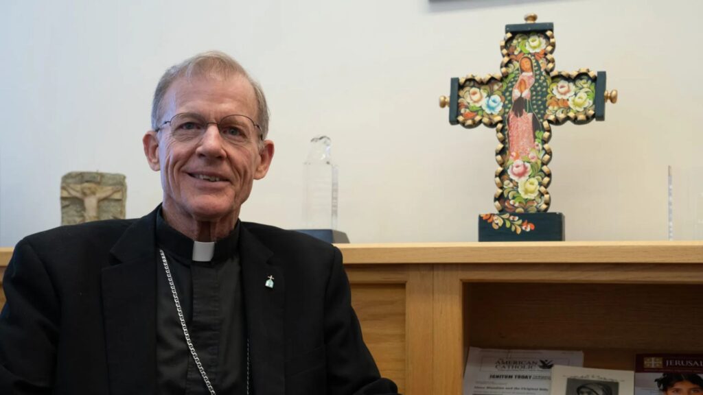 An elderly man in clerical attire sits in front of a bookshelf with a decorated cross and various books and objects in the background.