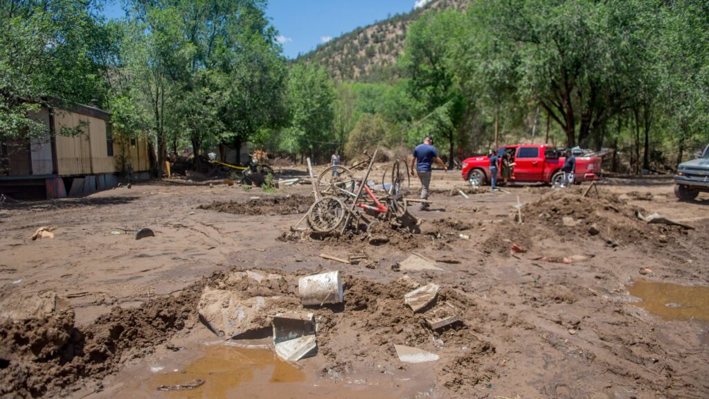 A muddy yard with scattered debris, a red truck, and two people working amid flood damage; trees and hills are visible in the background.