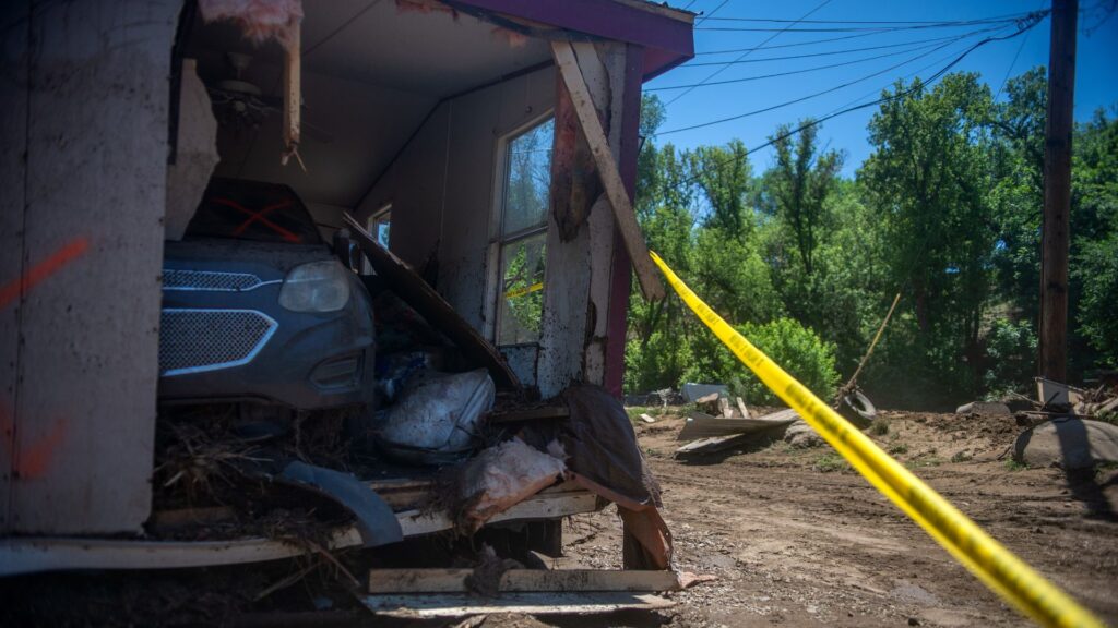 A car is lodged inside a damaged building, with debris and a yellow caution tape stretched across the scene; trees and dirt are visible in the background.