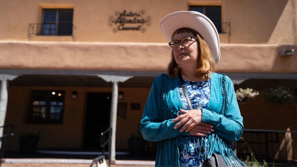 A woman in a white cowboy hat and teal fringe jacket stands in front of a tan adobe building with a sign reading "Hotel Chimayó de Santa Fe.