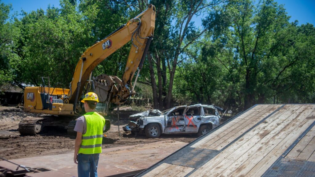 A construction worker observes an excavator lifting a damaged, spray-painted vehicle near a ramp in a wooded outdoor area.