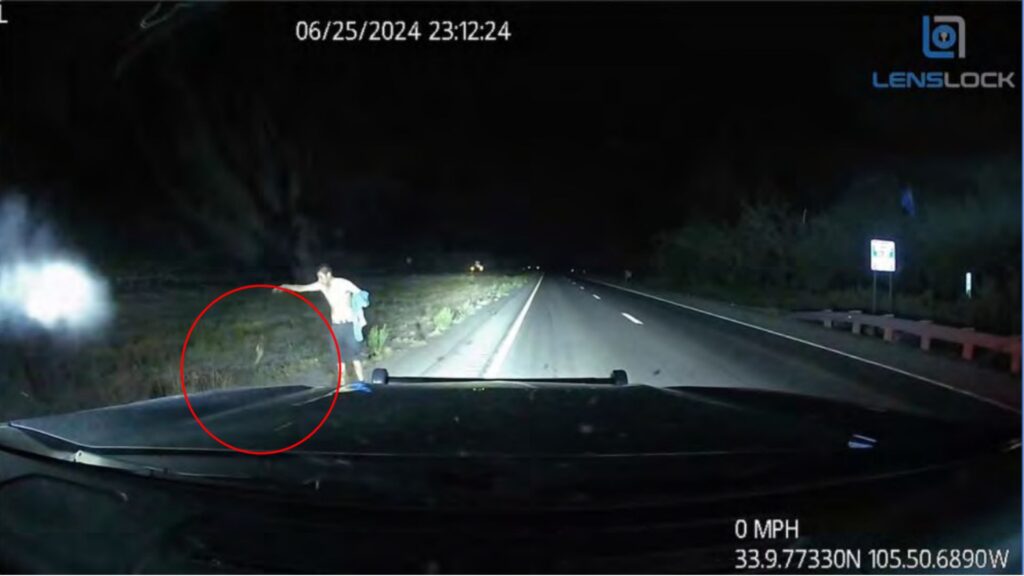 A person stands on the side of a dark, empty highway at night, illuminated by vehicle headlights; a timestamp and GPS coordinates are visible on the dashcam footage.