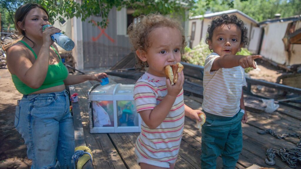 Two young children stand outdoors; one eats a sandwich while the other points. An adult woman drinks water nearby. The background shows makeshift housing and scattered belongings.