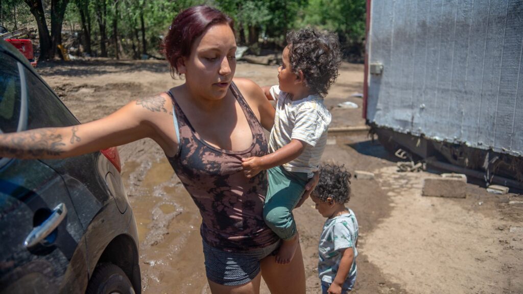 A woman stands outside next to a car, holding a young child while another child stands nearby on a muddy ground near a building.