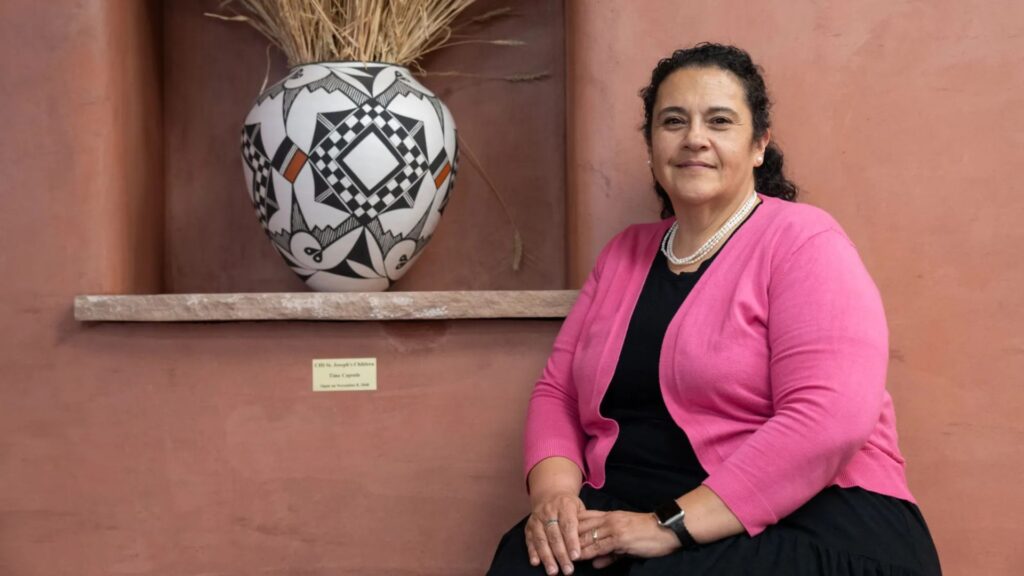 A woman in a pink cardigan sits beside a large patterned ceramic vase with dried grass, both placed against a terracotta-colored wall.