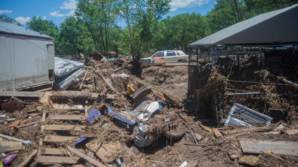 Debris and mud cover the ground between damaged structures and a car after a flood, with scattered wreckage and trees in the background under a blue sky.