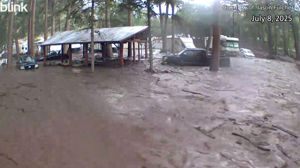 Floodwaters and mud surround a shelter, vehicles, and trees in a wooded area during a flood event. Date is July 8, 2025, with photo credit to Jason Fulcher.
