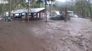 Floodwaters and mud surround a shelter, vehicles, and trees in a wooded area during a flood event. Date is July 8, 2025, with photo credit to Jason Fulcher.