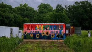 A colorful mural reading "Indigenous Farm Hub" with sunflowers and greenery, painted on a shipping container in a field with trees and grass.