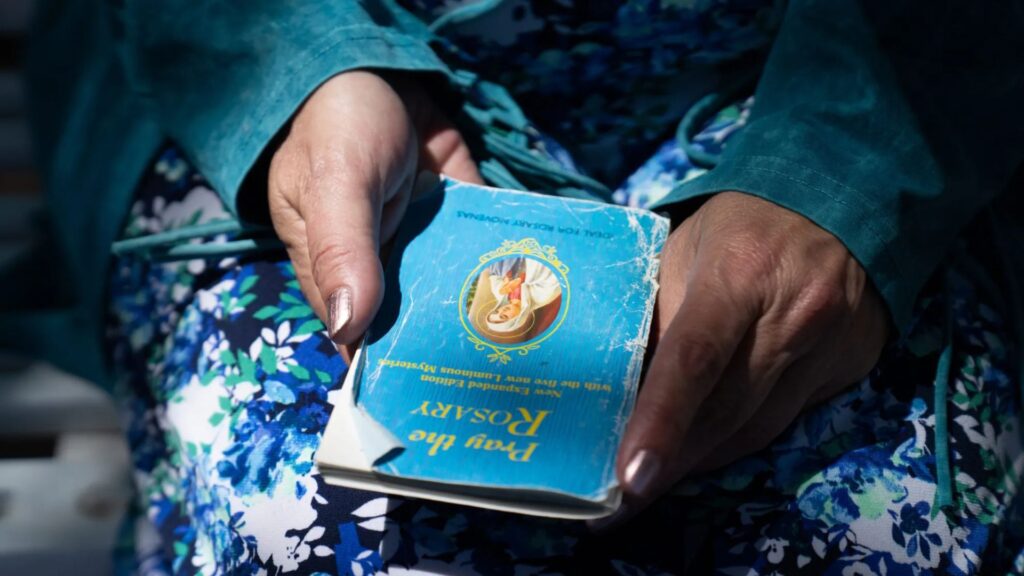 A person in a blue floral dress holds a worn booklet titled "Pray the Rosary" with an illustration of the Virgin Mary on the cover.