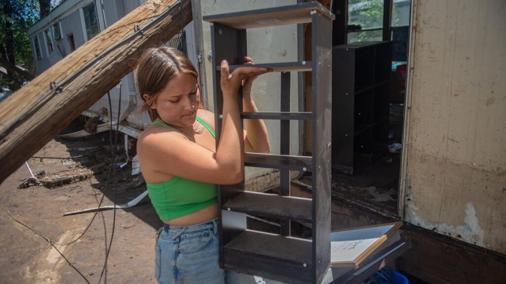 A woman in a green top and jeans carries a small shelving unit outside, near damaged buildings and debris.
