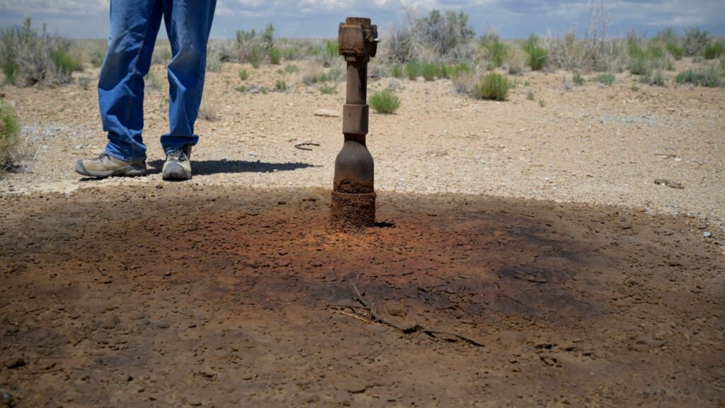 A rusted artillery shell is partially buried upright in dry, cracked ground, with a person in blue jeans and boots standing nearby. Sparse vegetation is visible in the background.